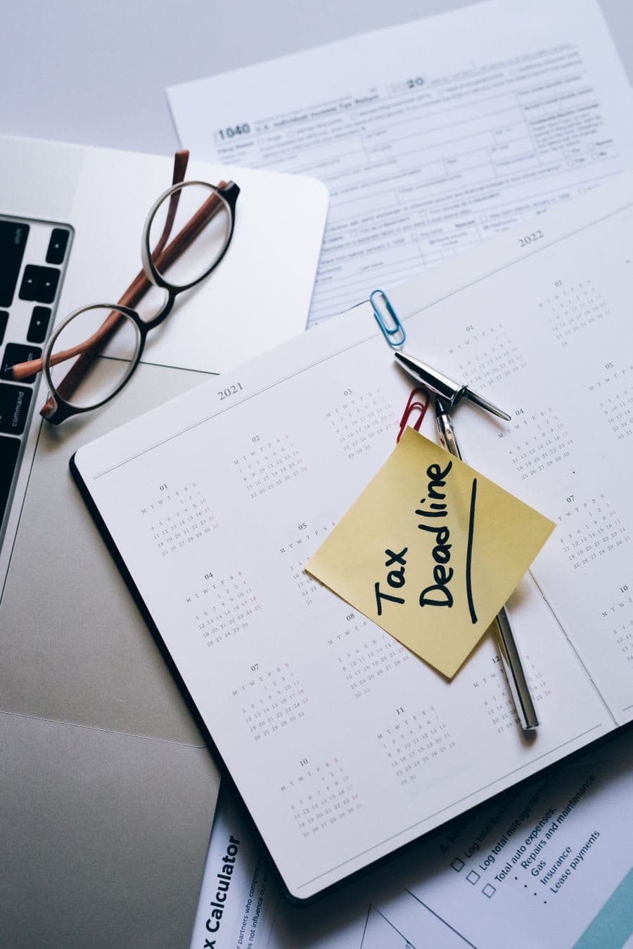 A tax deadline note stuck to a calendar beside forms and a laptop
