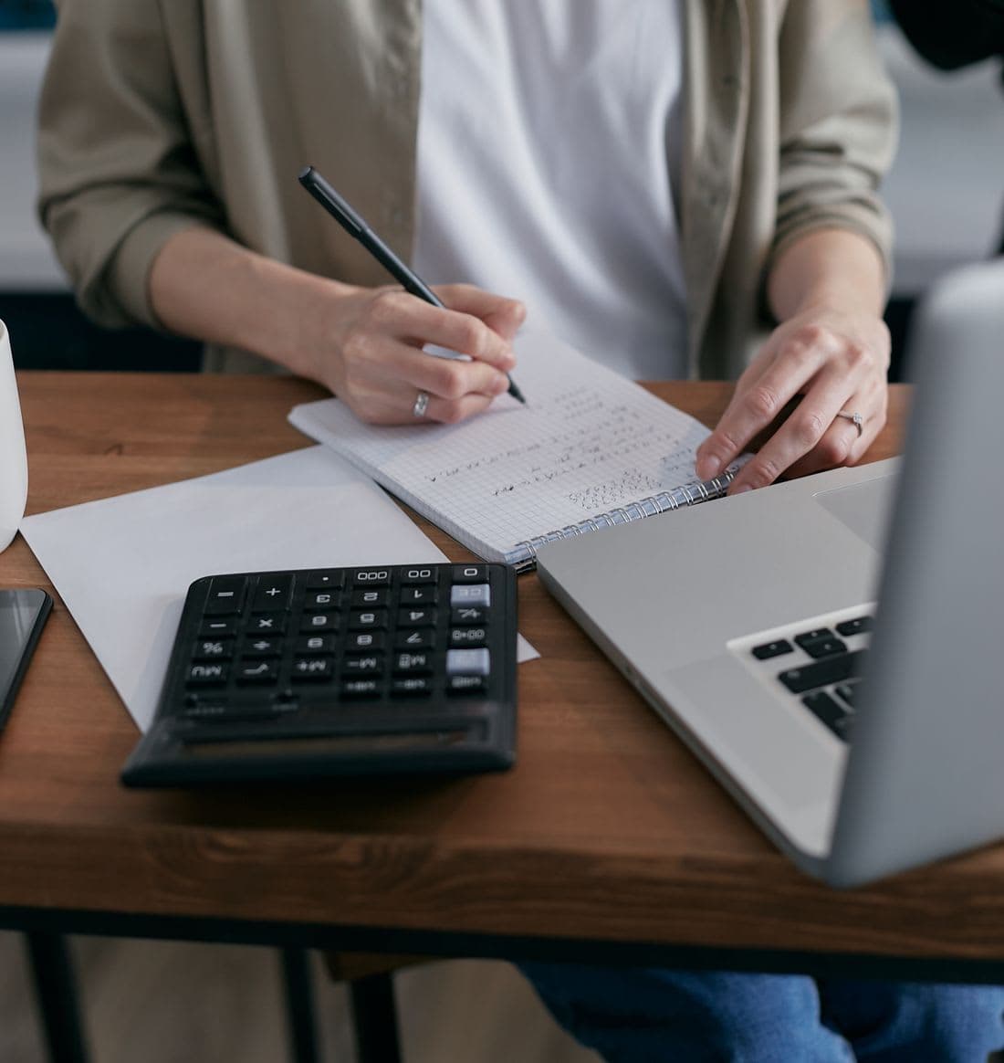 A bookkeeper writing calculations beside a laptop and calculator